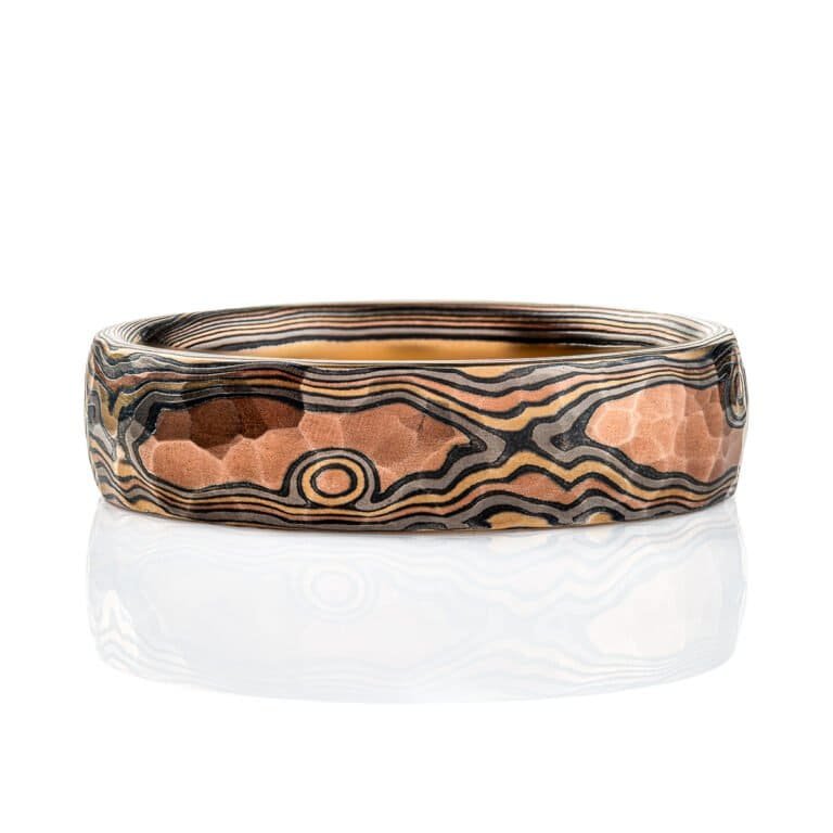 Mokume gane ring with a domed profile and textured surface all over, made with layers of red gold, yellow gold, palladium and oxidized silver (black), made in a twisting pattern that moves diagonally across the ring.