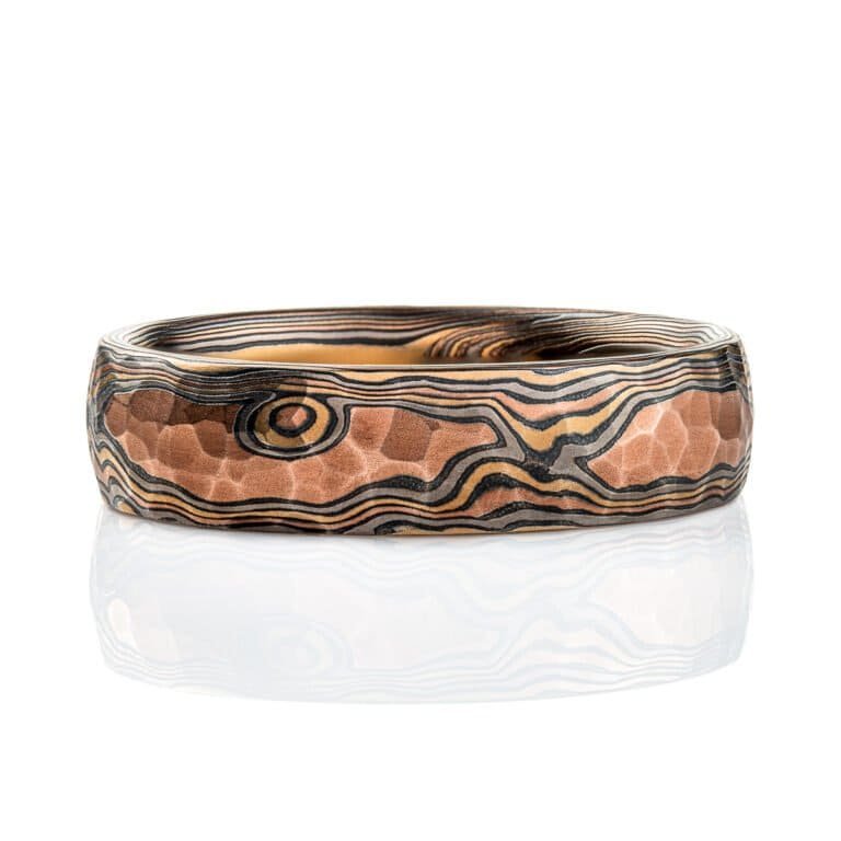 Mokume gane ring with a domed profile and textured surface all over, made with layers of red gold, yellow gold, palladium and oxidized silver (black), made in a twisting pattern that moves diagonally across the ring.