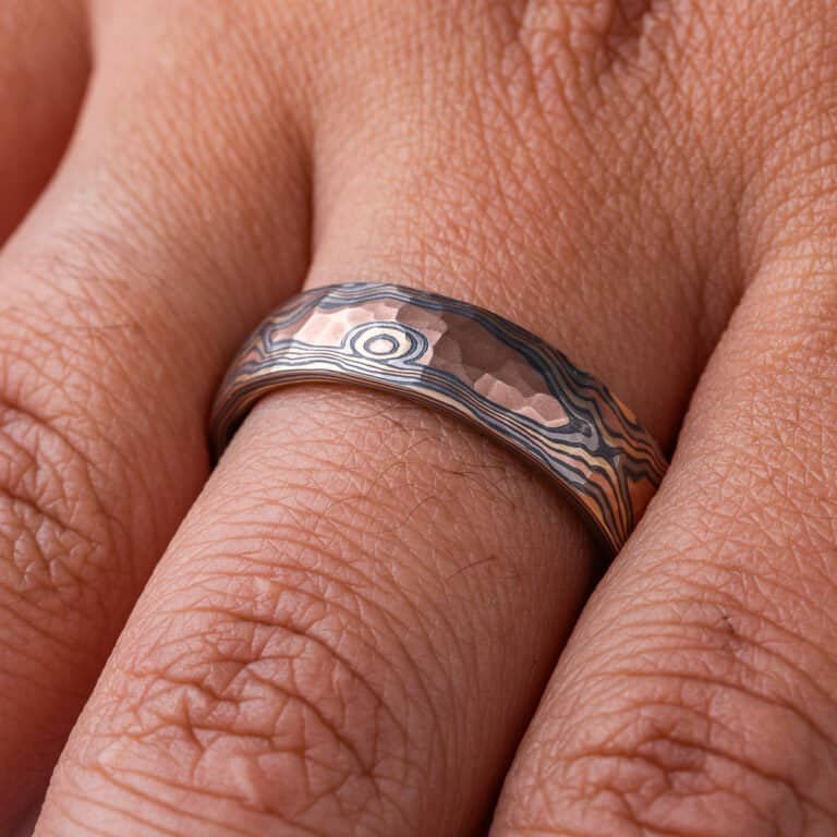 Mokume gane ring with a domed profile and textured surface all over, made with layers of red gold, yellow gold, palladium and oxidized silver (black), made in a twisting pattern that moves diagonally across the ring.