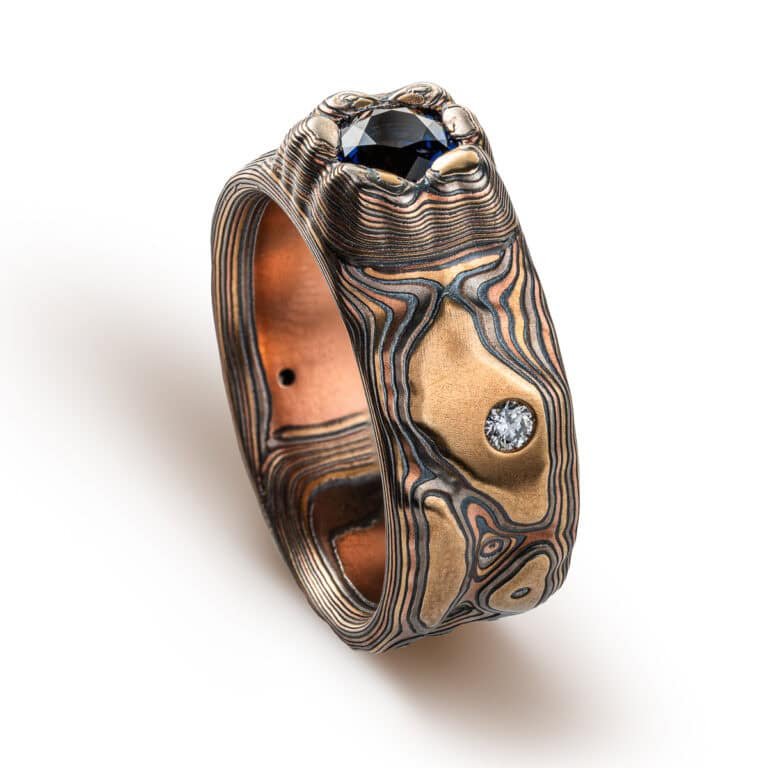 Large men's style mokume gane patterned ring, made to resemble rock formations and mountains, with a large dark blue and orange stone set into the ring in a raised area, intended to look like it's being excavated from the earth.