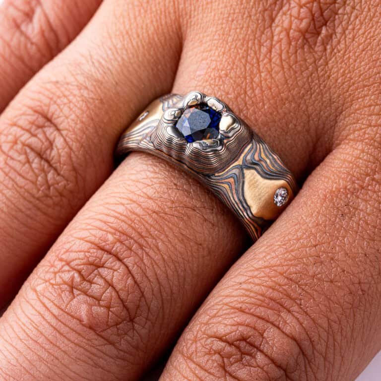 Large men's style mokume gane patterned ring, made to resemble rock formations and mountains, with a large dark blue and orange stone set into the ring in a raised area, intended to look like it's being excavated from the earth.