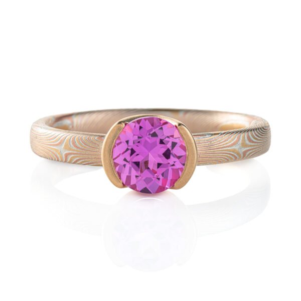 Narrow mokume gane patterned ring, subtle pattern, ring looks overall light yellow, and the ring has a larger round pink stone surrounded by solid yellow gold.