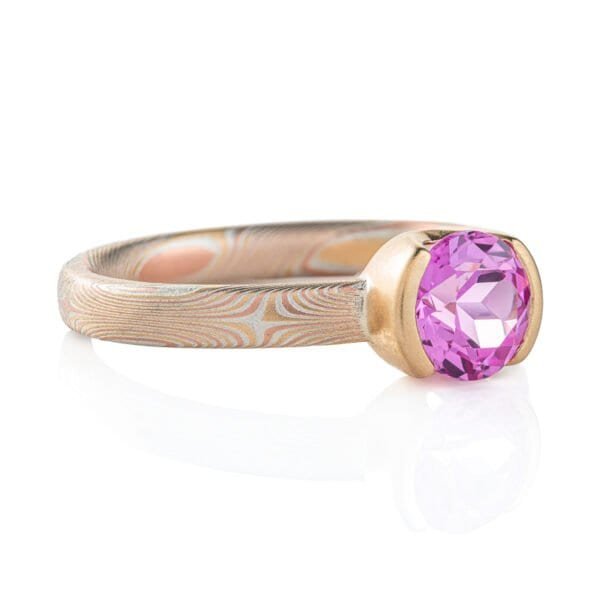 Narrow mokume gane patterned ring, subtle pattern, ring looks overall light yellow, and the ring has a larger round pink stone surrounded by solid yellow gold.