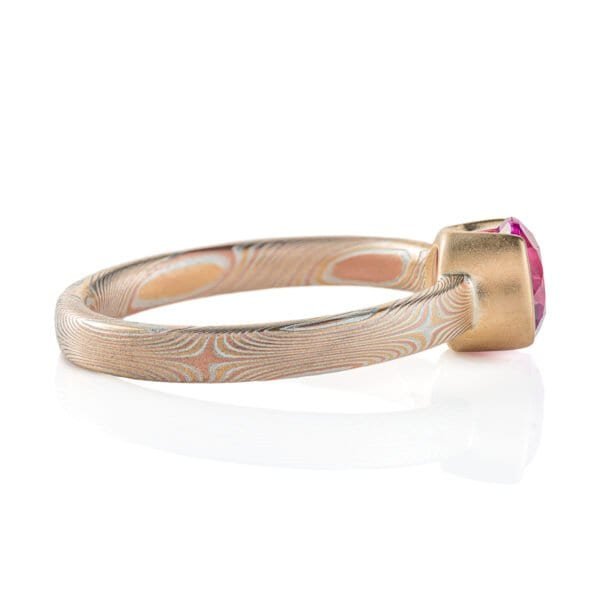 Narrow mokume gane patterned ring, subtle pattern, ring looks overall light yellow, and the ring has a larger round pink stone surrounded by solid yellow gold.