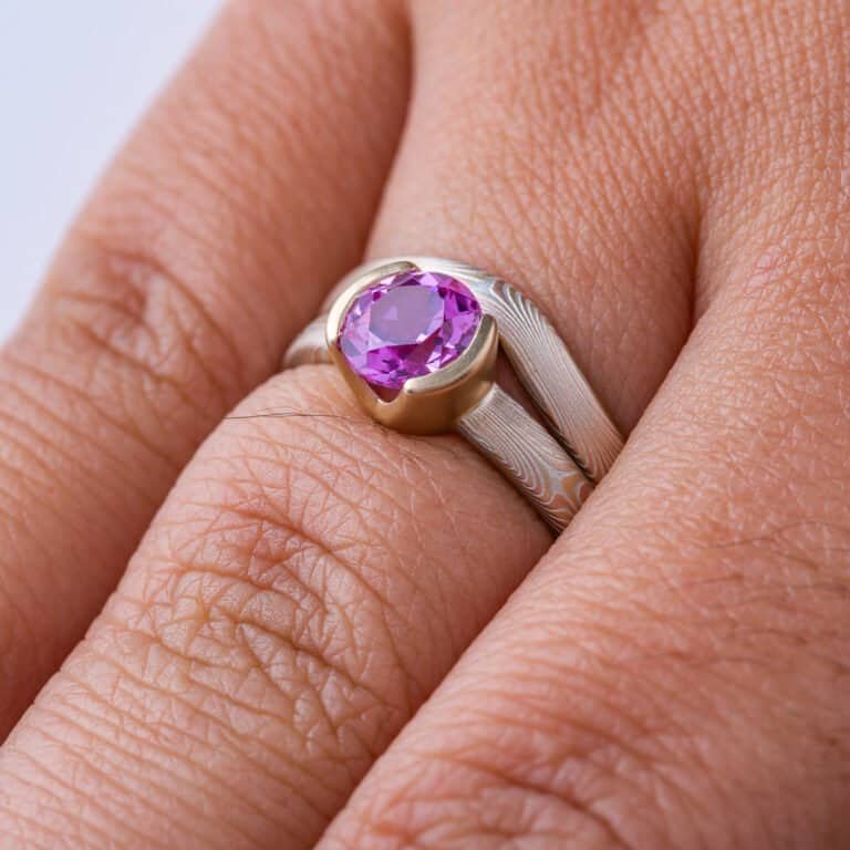 Pair of mokume gane patterned rings, one with a round pink stone, the other a band that curves around the stone and setting so the two can sit flush together. Overall warm light yellow gold look.