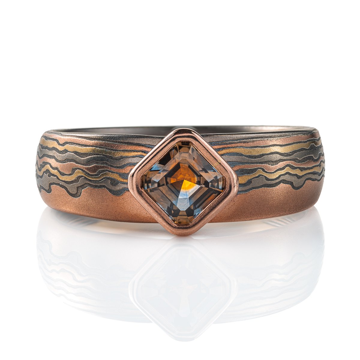 Mokume gane ring with a square stone set at an angle to look like a diamond, surrounded by a rim of red gold. Main ring is the same width as the stone at the front, and tapers towards the back, the pattern is made to look like thin clouds in a far away landscape.