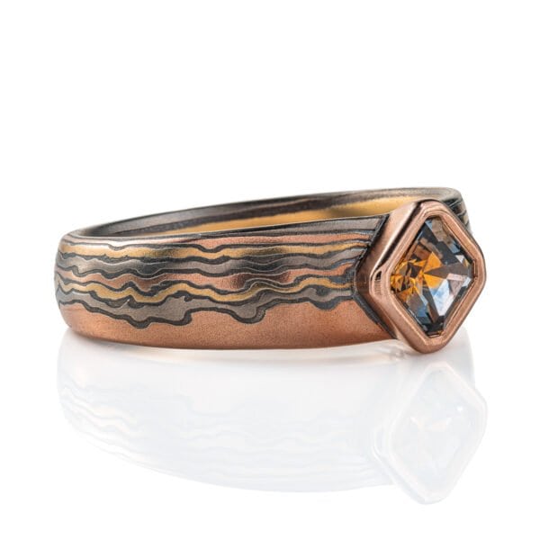 Mokume gane ring with a square stone set at an angle to look like a diamond, surrounded by a rim of red gold. Main ring is the same width as the stone at the front, and tapers towards the back, the pattern is made to look like thin clouds in a far away landscape.