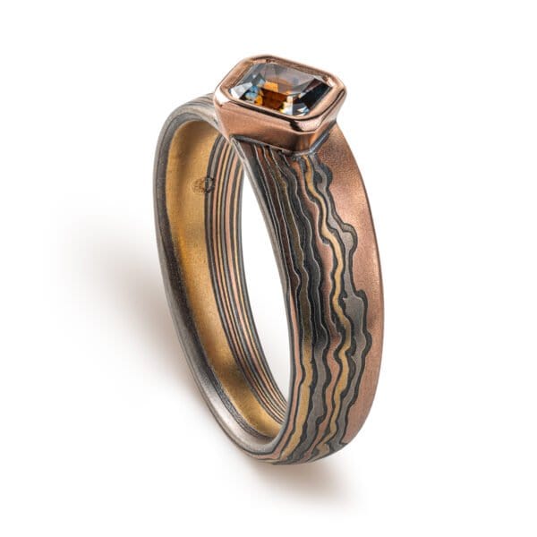 Mokume gane ring with a square stone set at an angle to look like a diamond, surrounded by a rim of red gold. Main ring is the same width as the stone at the front, and tapers towards the back, the pattern is made to look like thin clouds in a far away landscape.