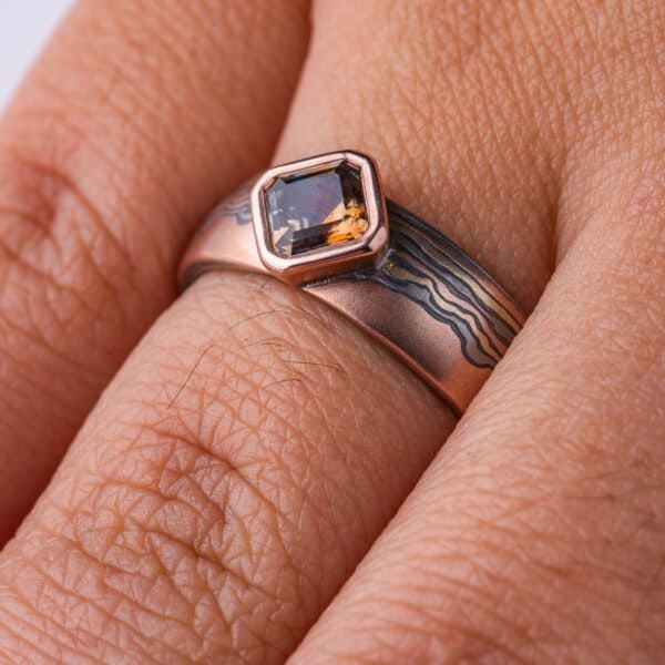 Mokume gane ring with a square stone set at an angle to look like a diamond, surrounded by a rim of red gold. Main ring is the same width as the stone at the front, and tapers towards the back, the pattern is made to look like thin clouds in a far away landscape.