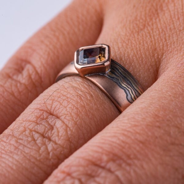 Mokume gane ring with a square stone set at an angle to look like a diamond, surrounded by a rim of red gold. Main ring is the same width as the stone at the front, and tapers towards the back, the pattern is made to look like thin clouds in a far away landscape.