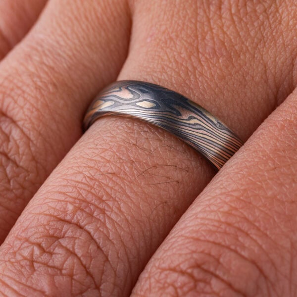 Mokume gane patterned ring, medium width, made in mixture of golds and dark gray metal in a twisting, swooping pattern.