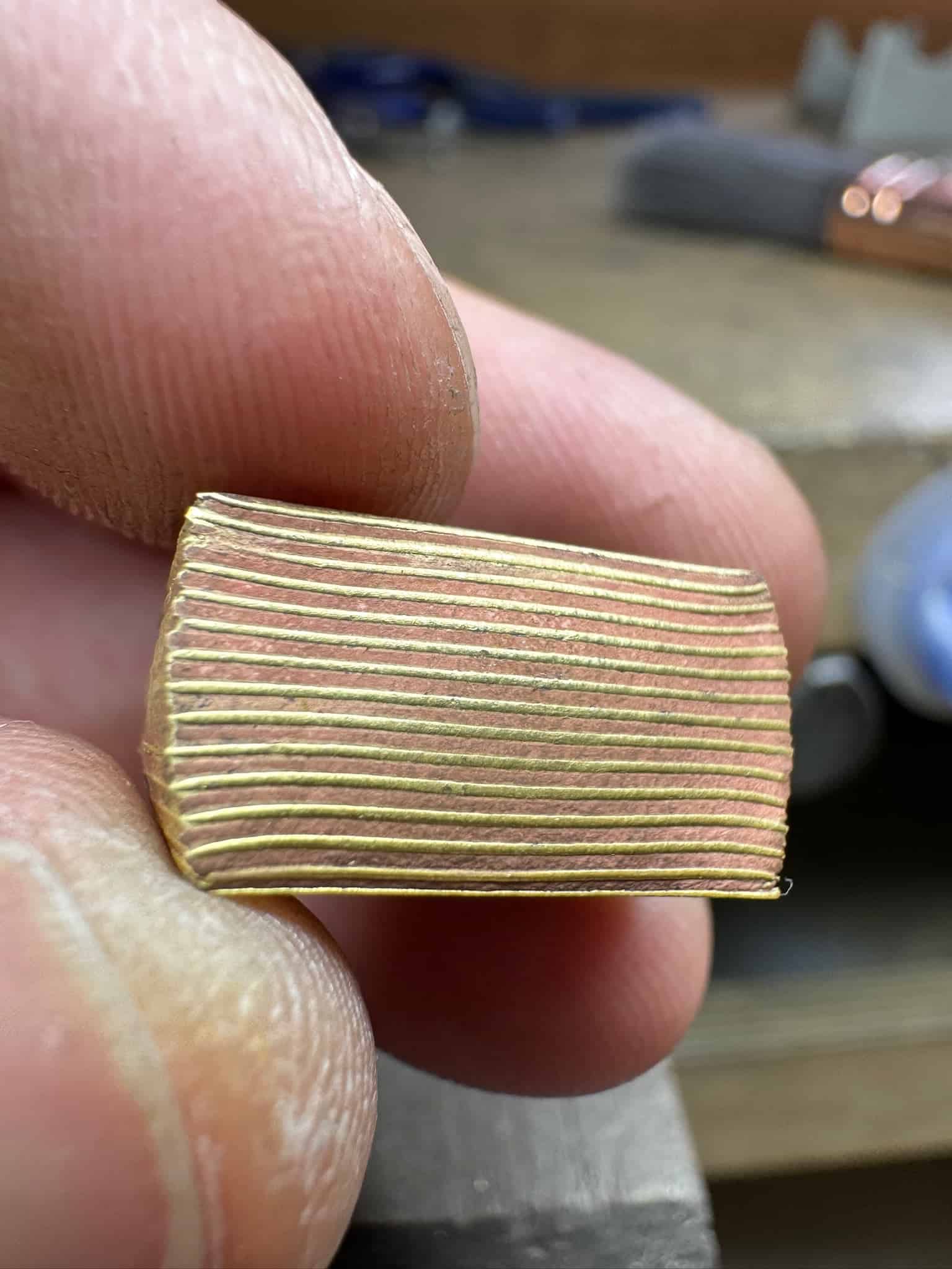 Close up of hand holding a fused metal billet - a stack of thin layers of metal that's been pressed together, alternating layers of yellow and red gold.