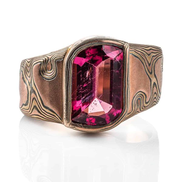 Mens mokume gane ring, with large pink stone surrounded by a border of mokume gane. The ring is wider at the top by the stone and tapers down towards the underside. Patterned in the woodgrain style, made with red gold, yellow gold and oxidized silver.