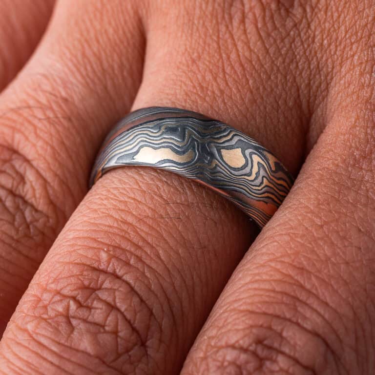 Wide men's style mokume gane band, patterned in twisting style, made with mainly yellow gold, palladium and oxidized silver, with one stripe of red gold.