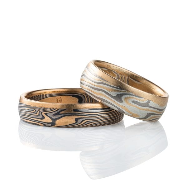 Pair of mokume gane rings, one lighter in color and wider, one darker in color and narrower, both made in a combination of yellow gold, palladium and silver, and each edged with yellow gold on one edge.