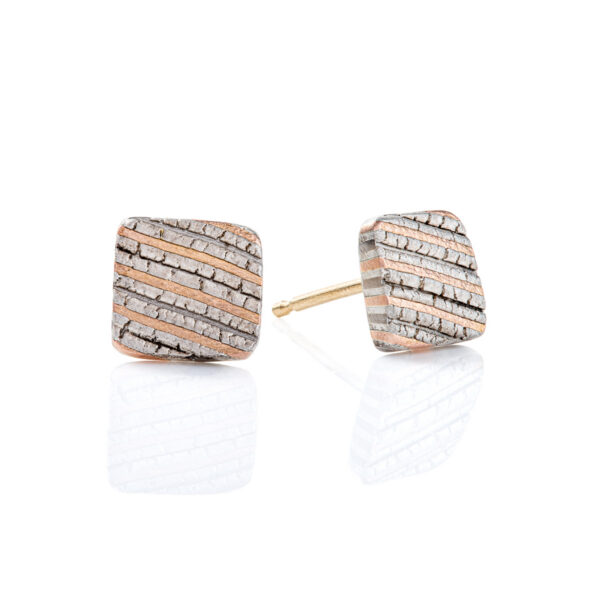 Small flat square stud earrings made with mokume gane, layered stripes of red gold, palladium and silver, with a crackly texture that resembles rock. .