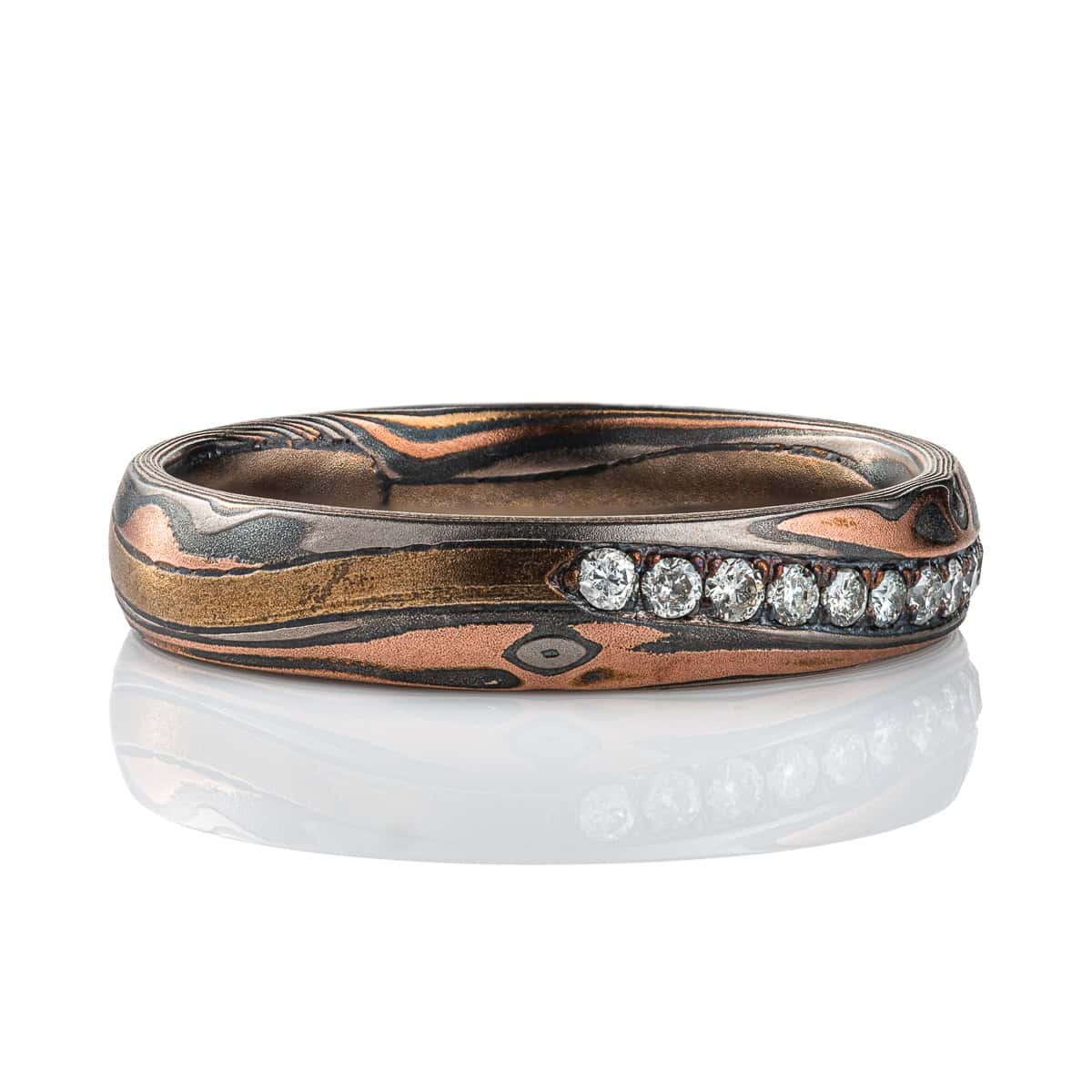Narrow mokume gane patterned band with undulating line of diamonds set flush into the surface. The band is made of warm toned metals in a woodgrain style pattern. .