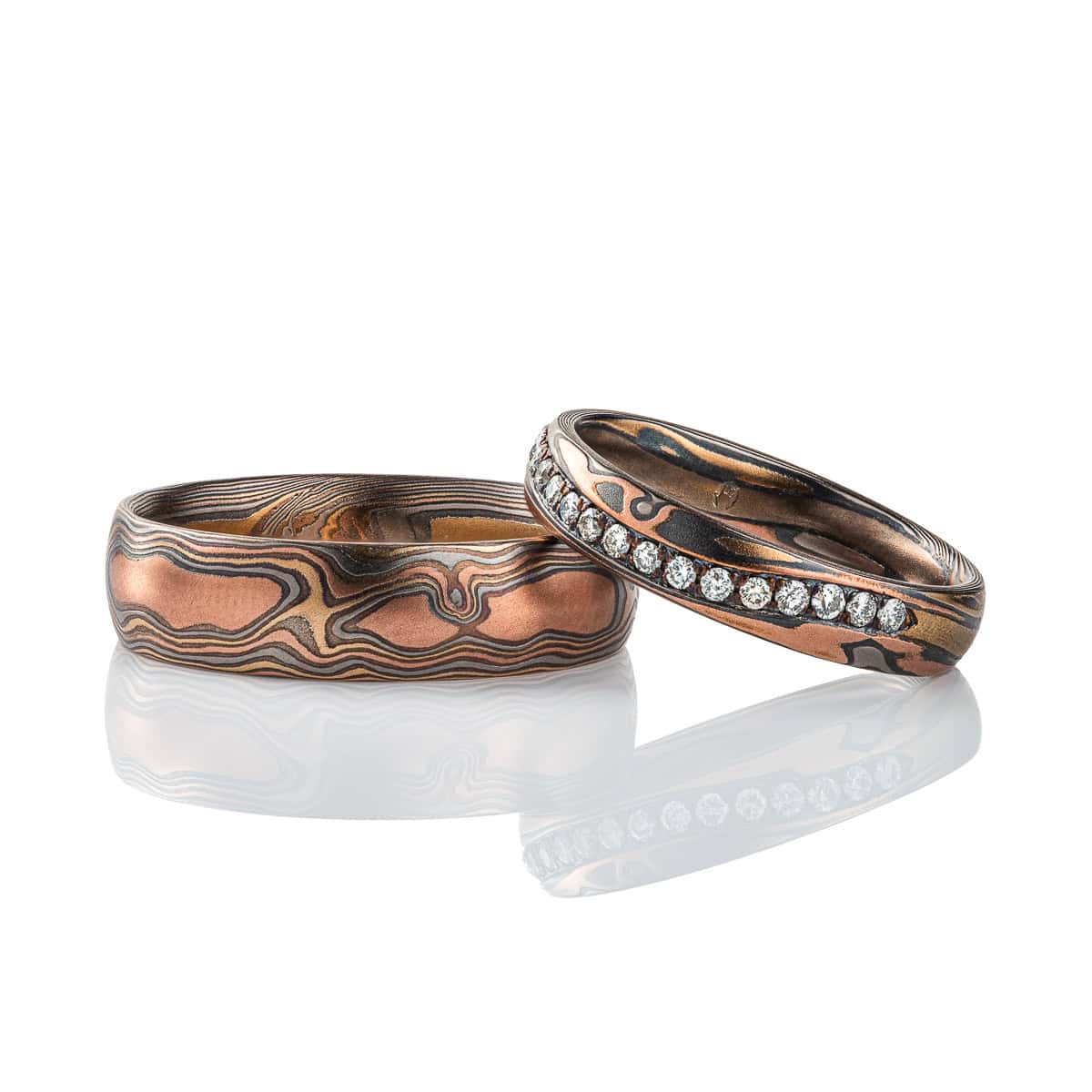 Pair of mokume gane patterned rings made in matching metal combination, with red gold, yellow gold, palladium and oxidized silver. The narrower ring also has a wavy line of small diamonds set into it.