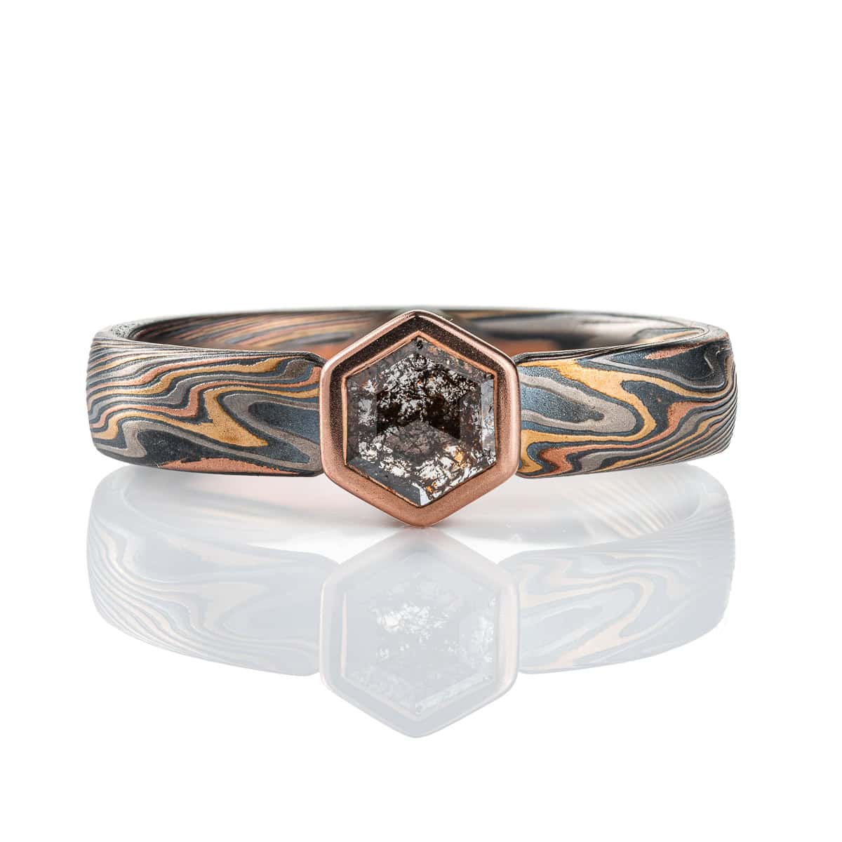 Mokume gane ring with a hexagonal stone held in a red gold bezel. The ring is made in a combination of red and yellow gold, palladium and silver. The stone is a salt and pepper diamond, with a speckled gray color.