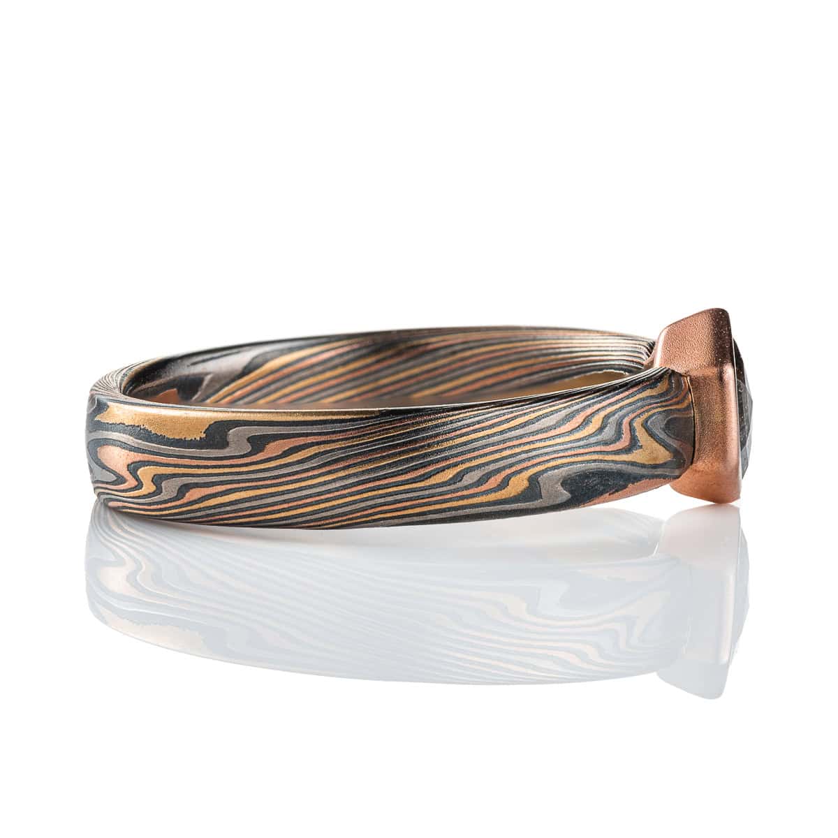 Mokume gane ring with a hexagonal stone held in a red gold bezel. The ring is made in a combination of red and yellow gold, palladium and silver. The stone is a salt and pepper diamond, with a speckled gray color.