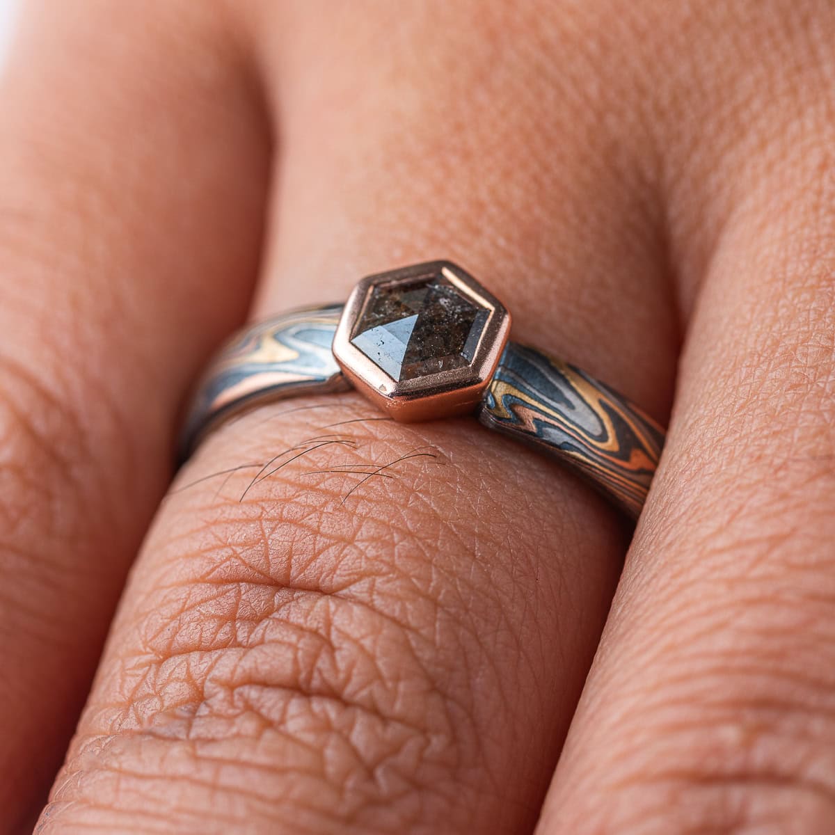 Mokume gane ring with a hexagonal stone held in a red gold bezel. The ring is made in a combination of red and yellow gold, palladium and silver. The stone is a salt and pepper diamond, with a speckled gray color.