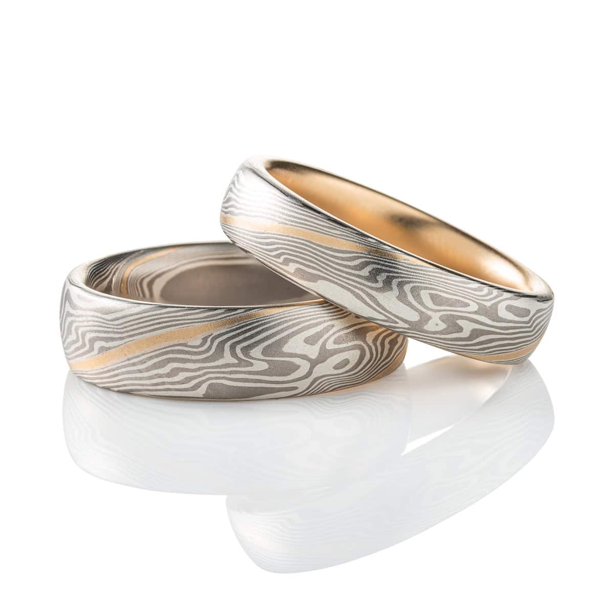 Pair of matching mokume gane patterned rings, each made in a silver and gray color palette with a single stripe of yellow gold. The smaller ring has also been lined with yellow gold.
