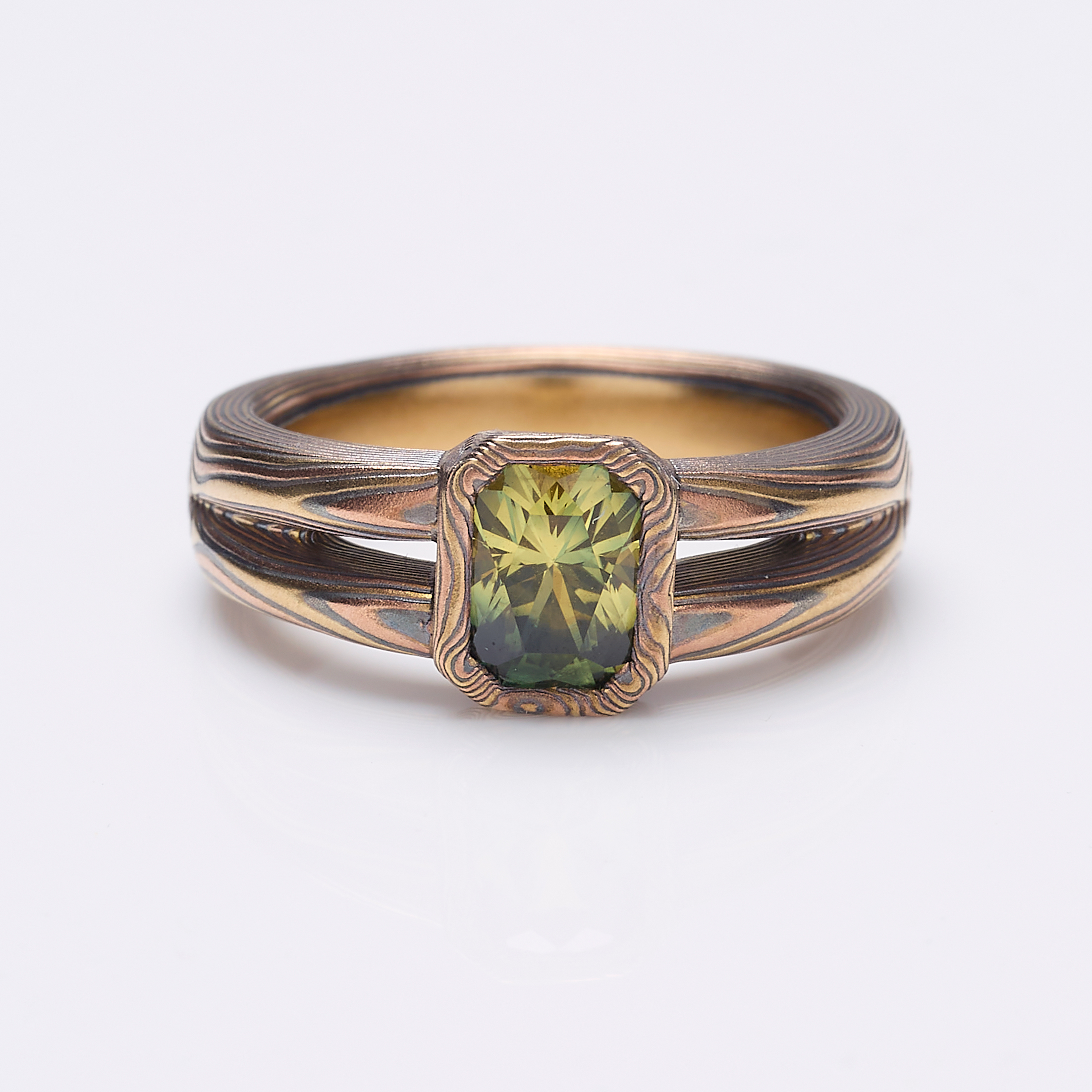 Mokume gane engagement ring with a split shank tapered towards the back of the ring, and a single green emerald cut sapphire placed at the front, with a rim of patterned metal around it.